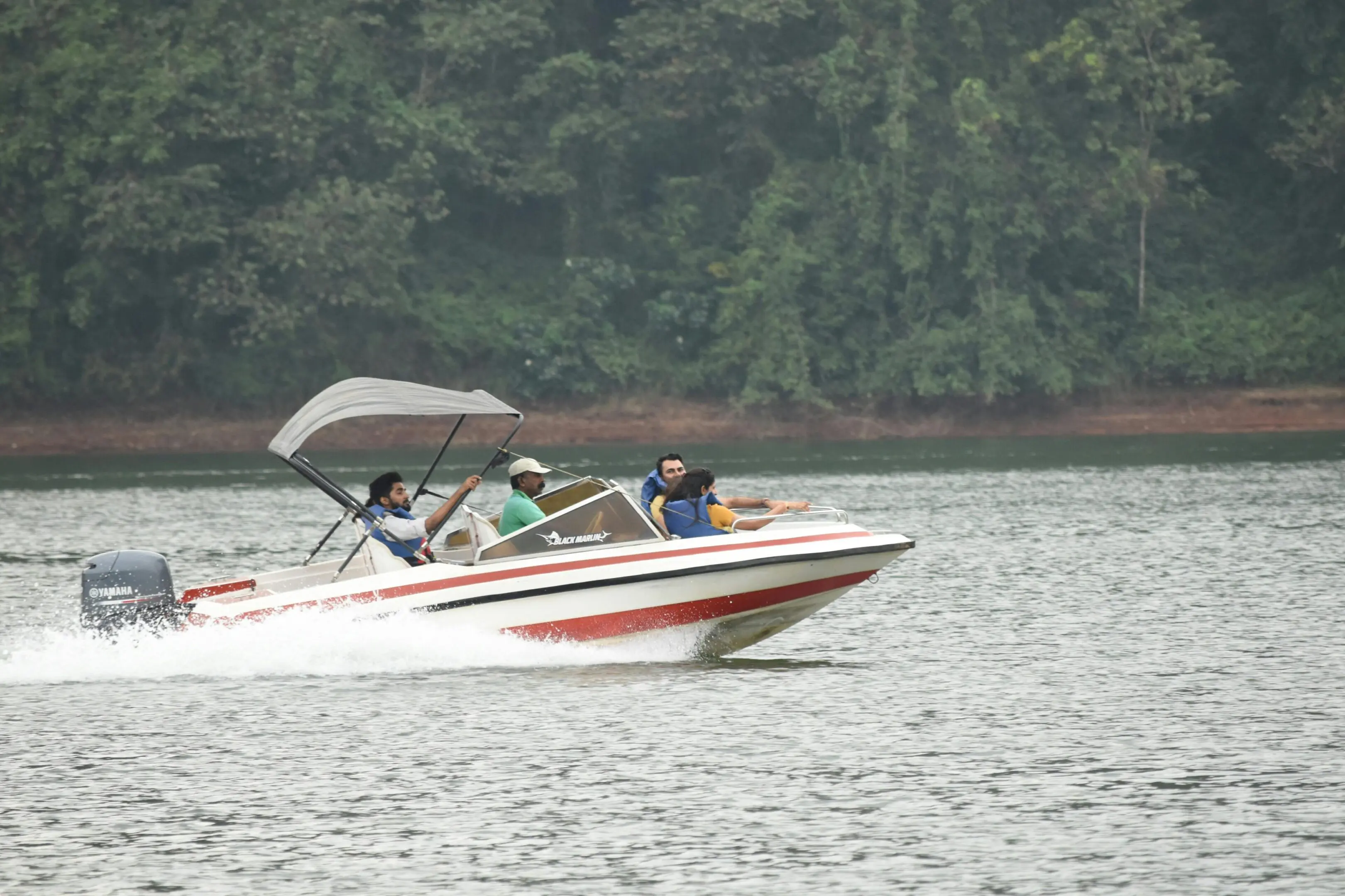 A speedboat with three people cruising on a lake near a forested shore.